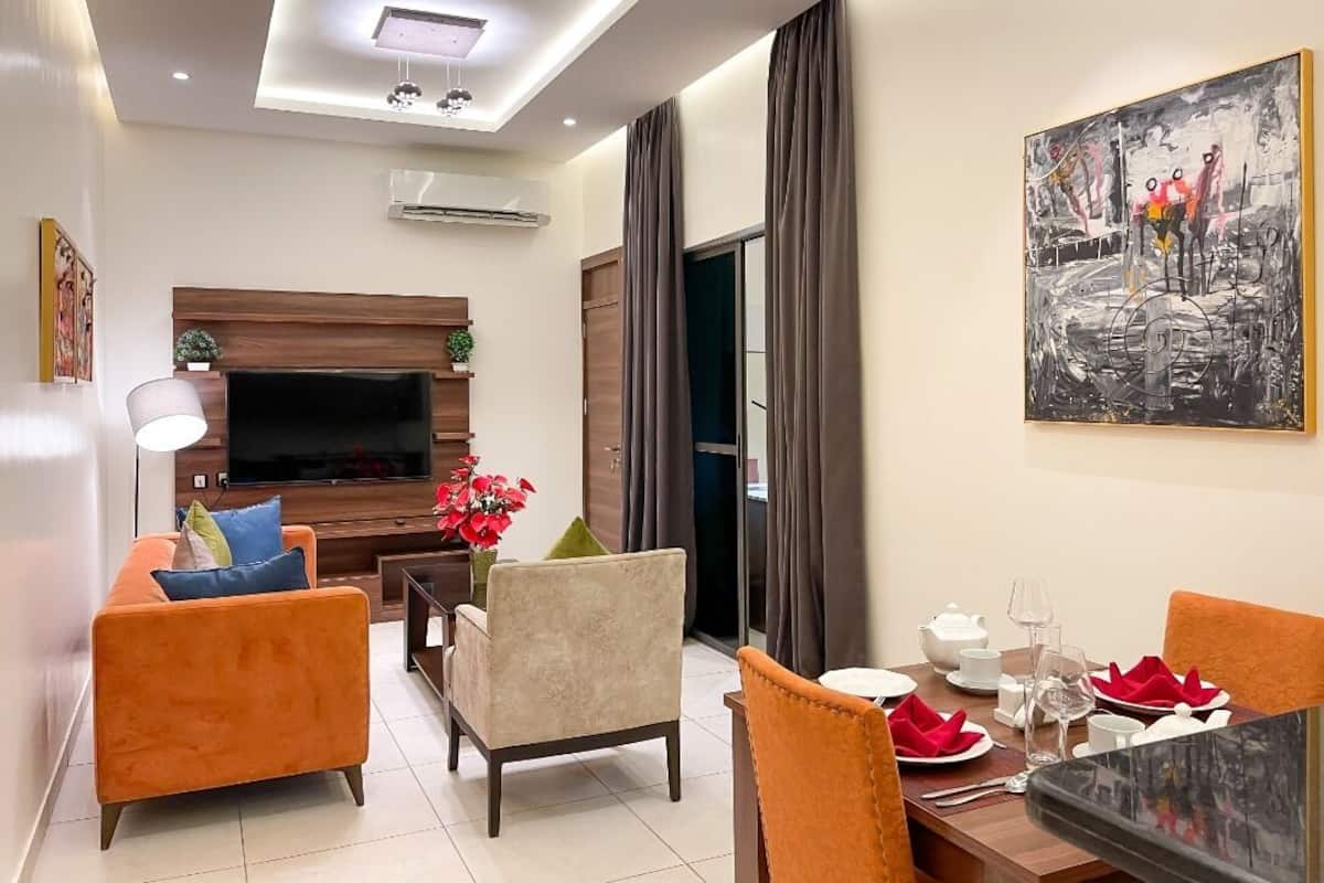 A modern living room with a flat-screen TV, orange sofas, a dining table set for two, and a large abstract painting on the wall.