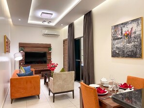 A modern living room with a flat-screen TV, orange sofas, a dining table set for two, and a large abstract painting on the wall. - OMEDEL LUXURY (Enugu)