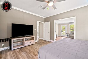 A bedroom with a bed, a TV on a wooden stand, a ceiling fan, and a door leading to a balcony.