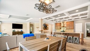 A modern living room with a wooden dining table, grey sofa, and a kitchen area with wooden cabinets and a gas stove.