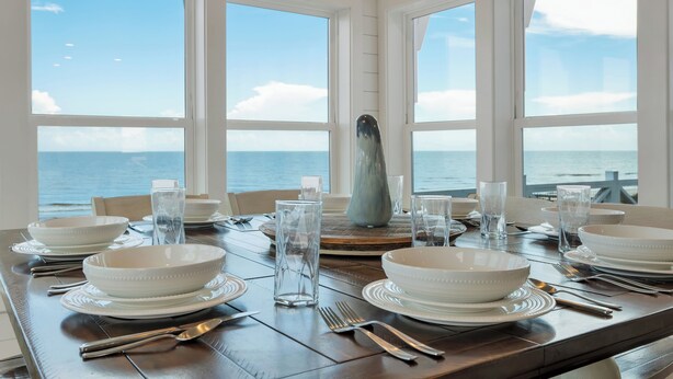 A dining table set with white plates, bowls, and cutlery, overlooking the sea. - Island Rhythm: Beachfront, Community Pool, Hot Tub, Pet Friendly! (St. George Island)