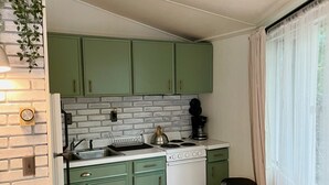 Una cocina compacta con gabinetes verdes, una estufa blanca y un lavamanos. Hay un bote de basura pequeño y una ventana con cortinas.