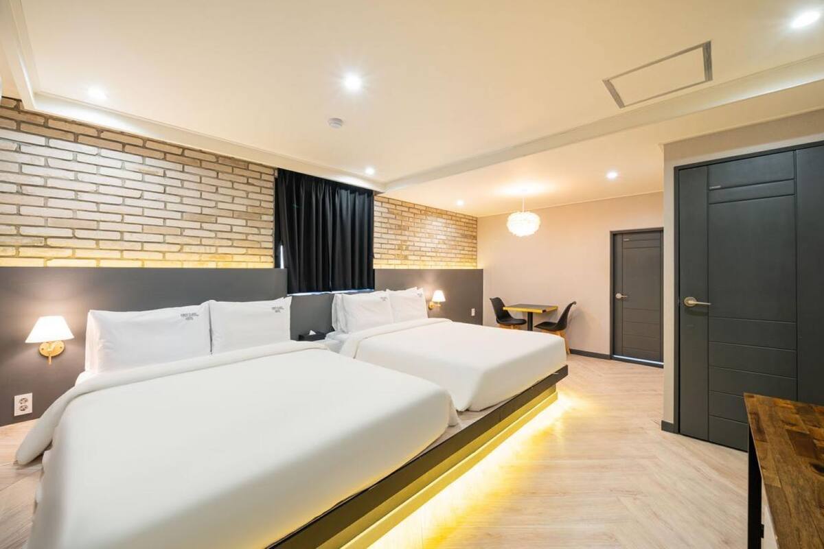 A hotel room with two beds, a brick accent wall, a dark door, and a small table with chairs.