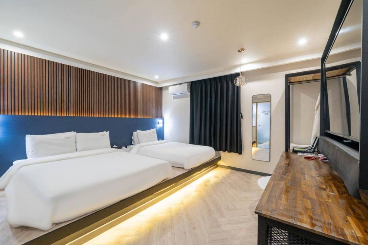 A hotel room with two beds, a wooden desk, a mirror, and a window with curtains.