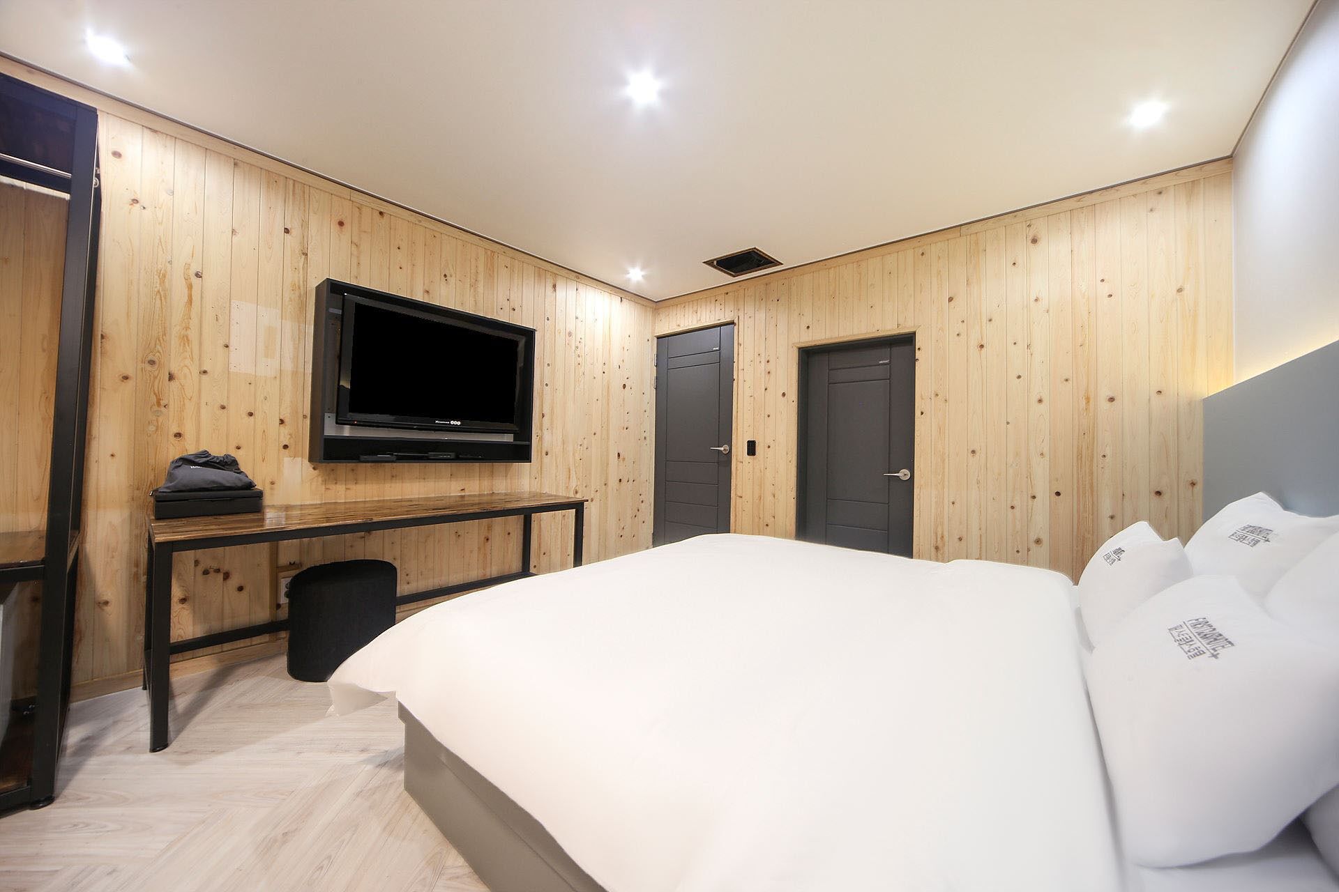 Een moderne hotelkamer met houten paneelwanden, een flatscreen televisie aan de muur, een bed met witte lakens, een klein bureau met stoel en een deur.