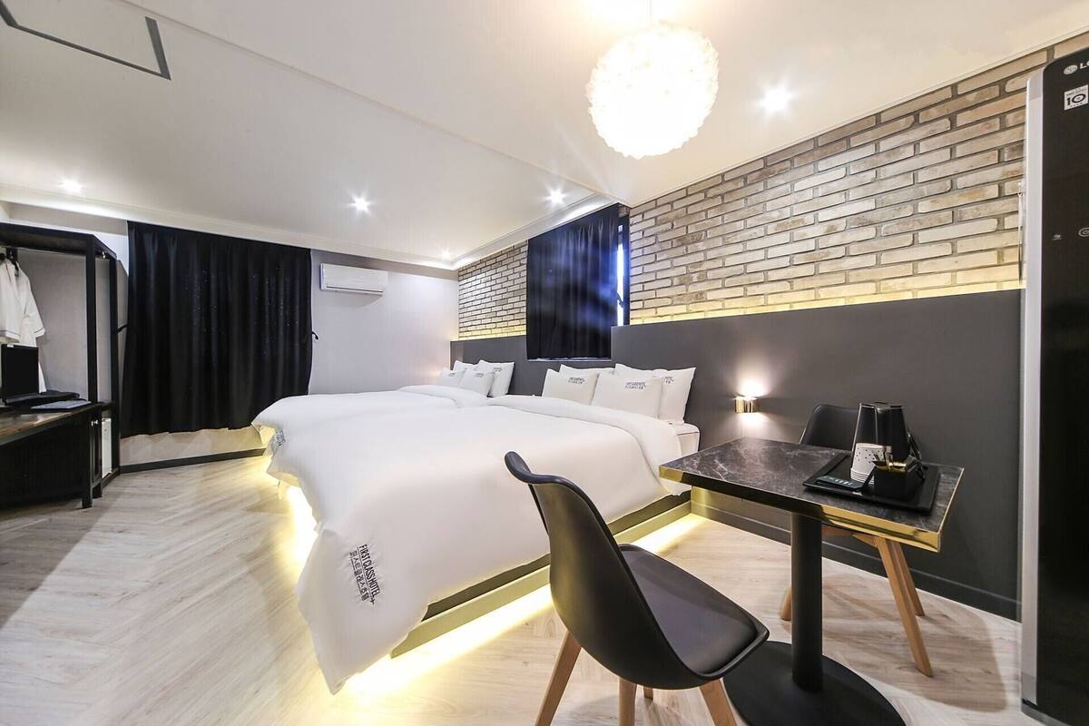 A modern hotel room with a large bed, a desk with a chair, a small table with a lamp, and a brick accent wall.
