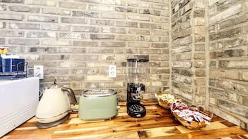 A kitchen with a brick wall, a kettle, a toaster, a blender, and a basket of bread.