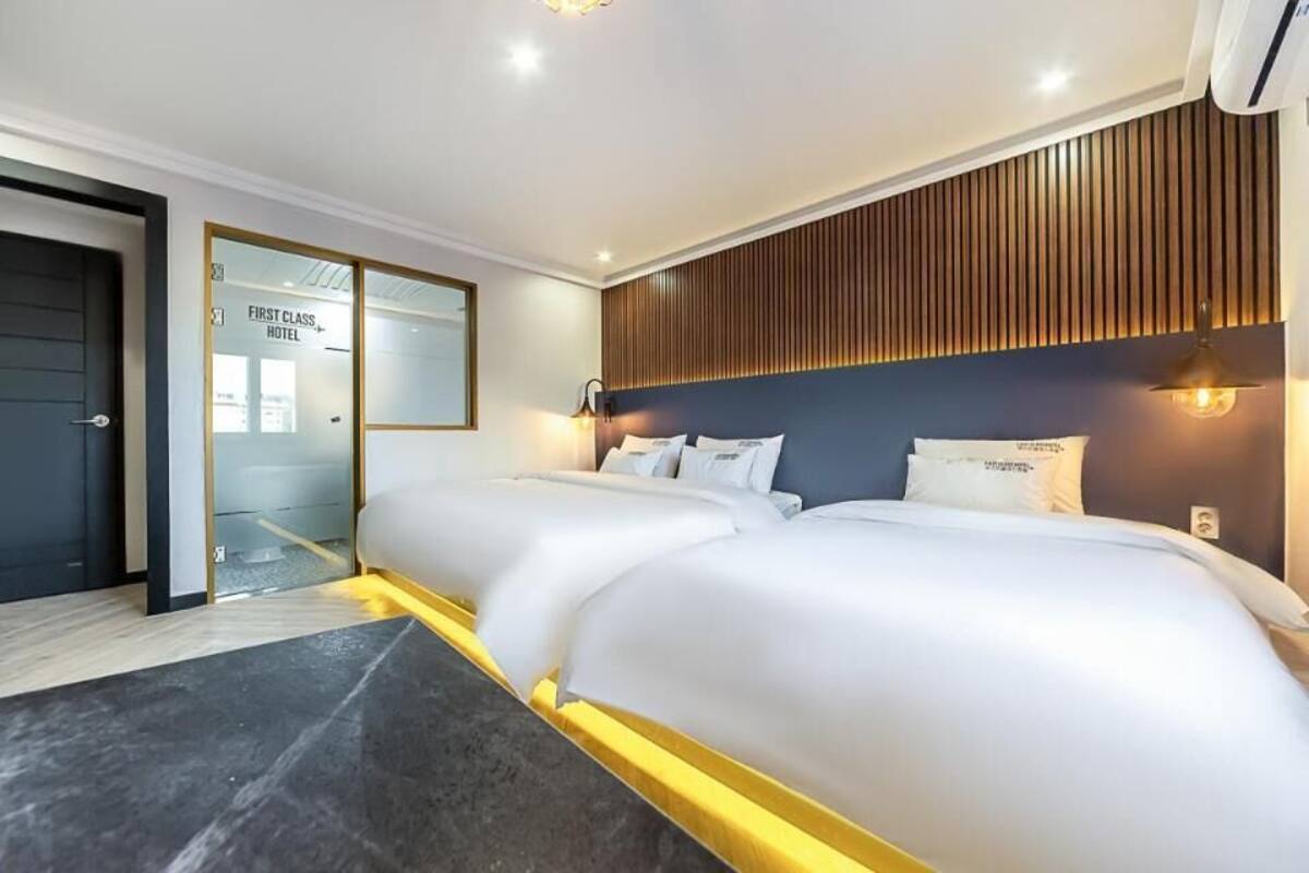 A hotel room with a bed, a bedside table, a wall-mounted light, and a door leading to another room.