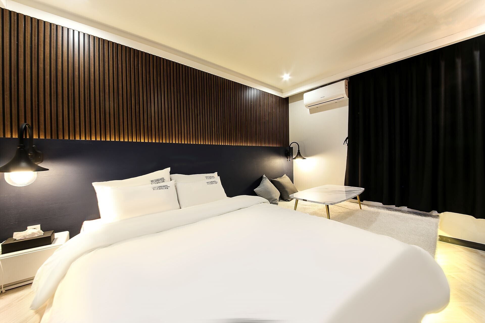 A modern hotel room with a large bed, bedside lamps, a small table, and a black curtain.