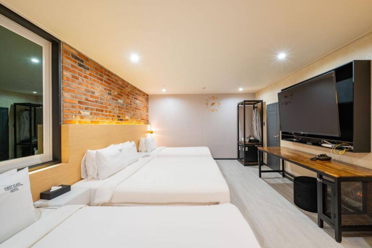 A hotel room with two beds, a TV, a wooden bench, and a brick wall.