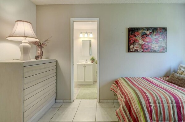A bedroom with a bed, a bedside lamp, a painting, and a bathroom visible through an open door.