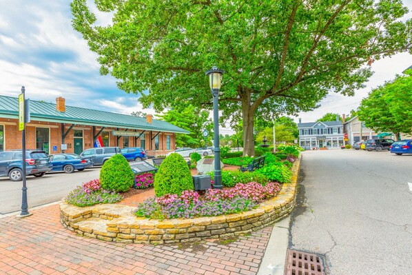 A street with a brick sidewalk, a stone planter with flowers, a lamp post, and a building with a green roof.