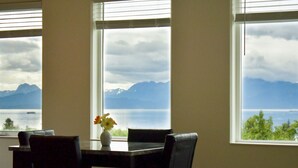 Ein Essbereich mit Tisch und Stühlen, einer Vase mit Blumen und Blick auf einen See und Berge durch große Fenster.