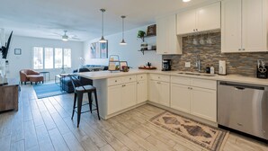 Una cocina moderna con armarios blancos, lavavajillas integrado y salpicadero de piedra. Hay una zona de bar con taburetes y un espacio para preparar café.