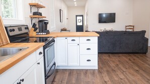 Une cuisine moderne avec des meubles blancs, un plan de travail en bois et un four encastré. Le salon est équipé d’un canapé gris et d’un téléviseur à écran plat.