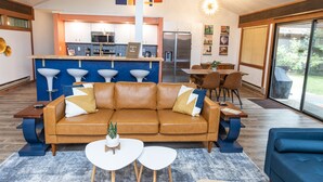 Un salon moderne avec un canapé en cuir marron, une table basse blanche et un fauteuil bleu. On aperçoit, au fond, une cuisine avec un coin bar.