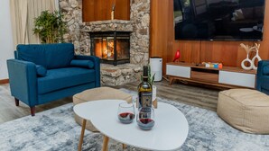 Un salon doté d’une cheminée en pierre, d’un canapé bleu, d’une table basse blanche sur laquelle sont posés des verres à vin, et d’un téléviseur à écran plat fixé au mur.