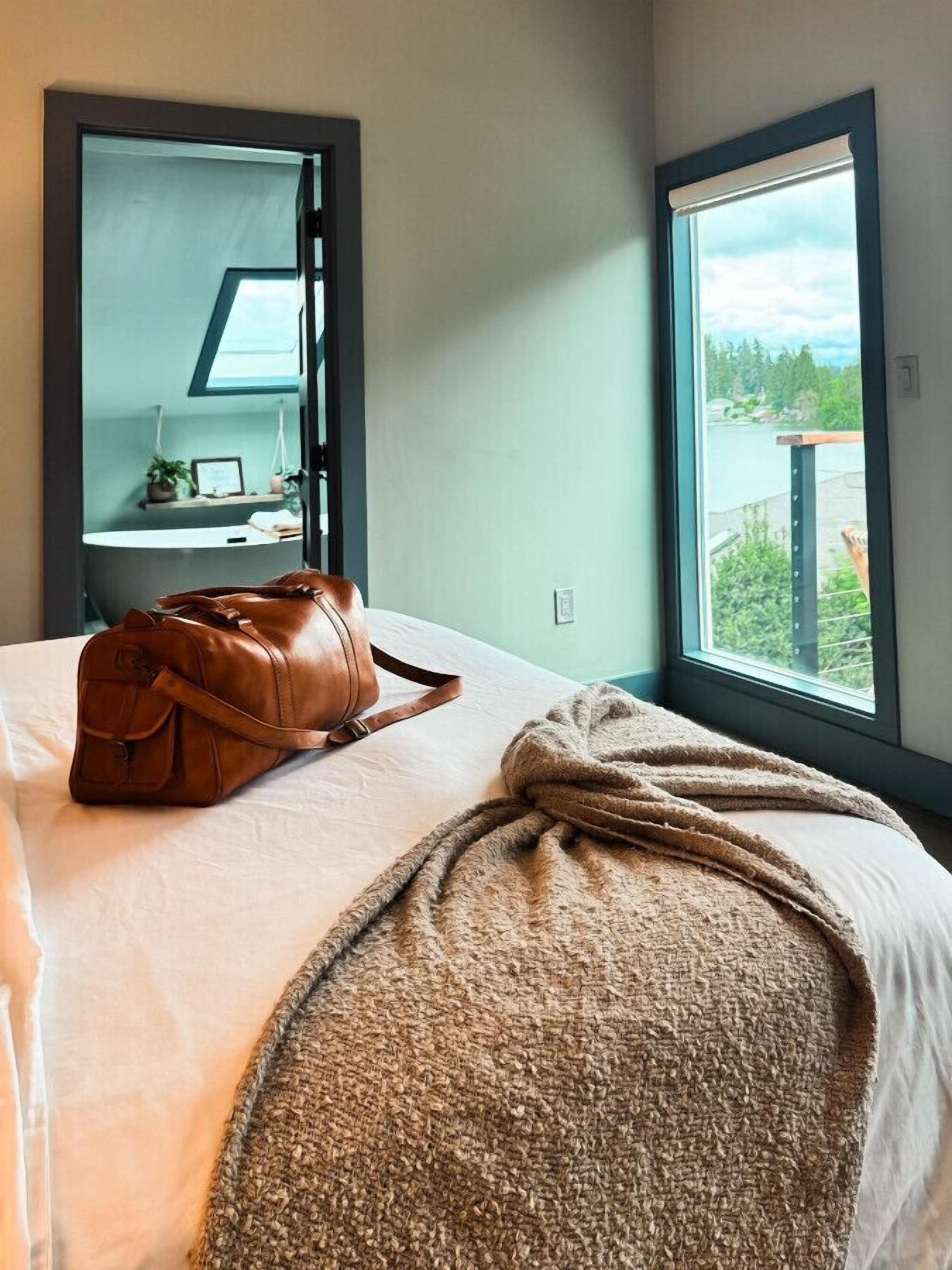 A hotel room with a bed, a brown leather bag, a window with a view of greenery, and a mirror reflecting a bathroom.