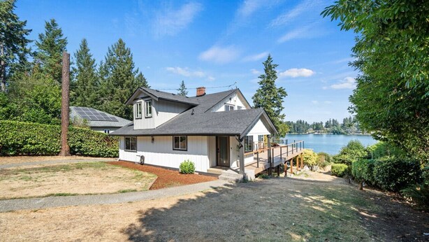 A two-story house with a deck overlooking a lake, surrounded by trees and a well-maintained lawn.