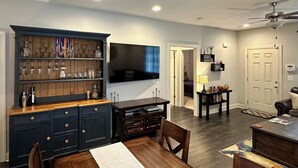 A living room with a dark wooden entertainment center, a flat-screen TV, a dining table with chairs, and a ceiling fan. - Auburn Gameday Townhome (Opelika)