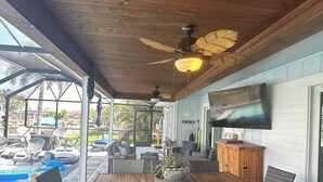 Une terrasse extérieure avec une table en bois et des chaises en osier, un ventilateur de plafond et un téléviseur fixé au mur.