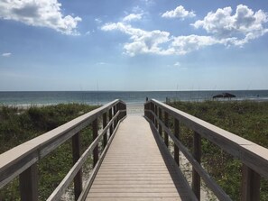 Ein hölzerner Steg führt zu einem Strand mit blauem Himmel und weiter Ferne.