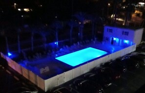Ein rechteckiger Pool mit blau beleuchtetem Rand, umgeben von einem Zaun und Palmen.