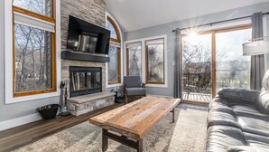 Un salon moderne doté d’une cheminée en pierre, d’une table basse en bois, d’un canapé en cuir et de grandes fenêtres offrant une vue sur l’extérieur.