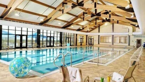Une piscine intérieure dotée d’un haut plafond et de poutres en bois, entourée de chaises longues et d’une grande fenêtre offrant une vue sur le paysage.