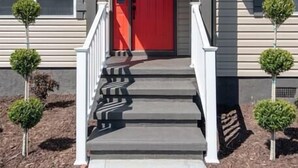 Une maison avec une porte rouge, le numéro « 1485 » au-dessus de l’entrée et un petit porche avec des marches menant à la porte.