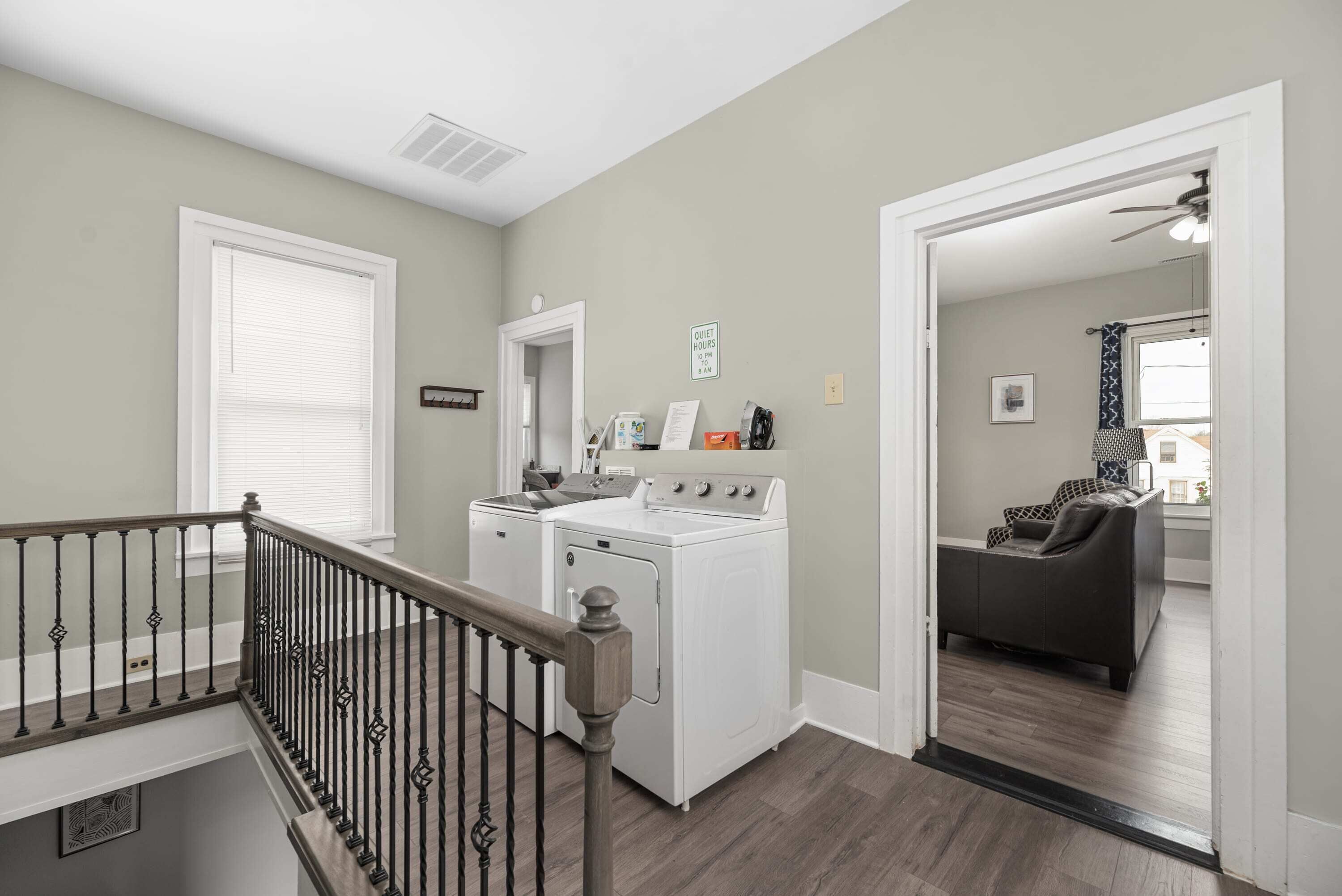 Un espace buanderie équipé d’un lave-linge et d’un sèche-linge, un escalier avec une rampe et une porte donnant sur un salon avec un canapé.