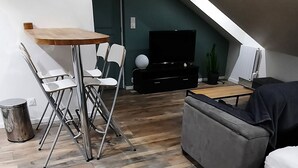 Et moderne loftrum med skrå vægge, trægulv, et spisebord med stole, en grå sofa og et fjernsyn.