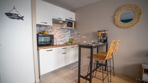 Cozinha compacta com armários brancos, micro-ondas e máquina de café. Há duas cadeiras de vime e uma mesa pequena.