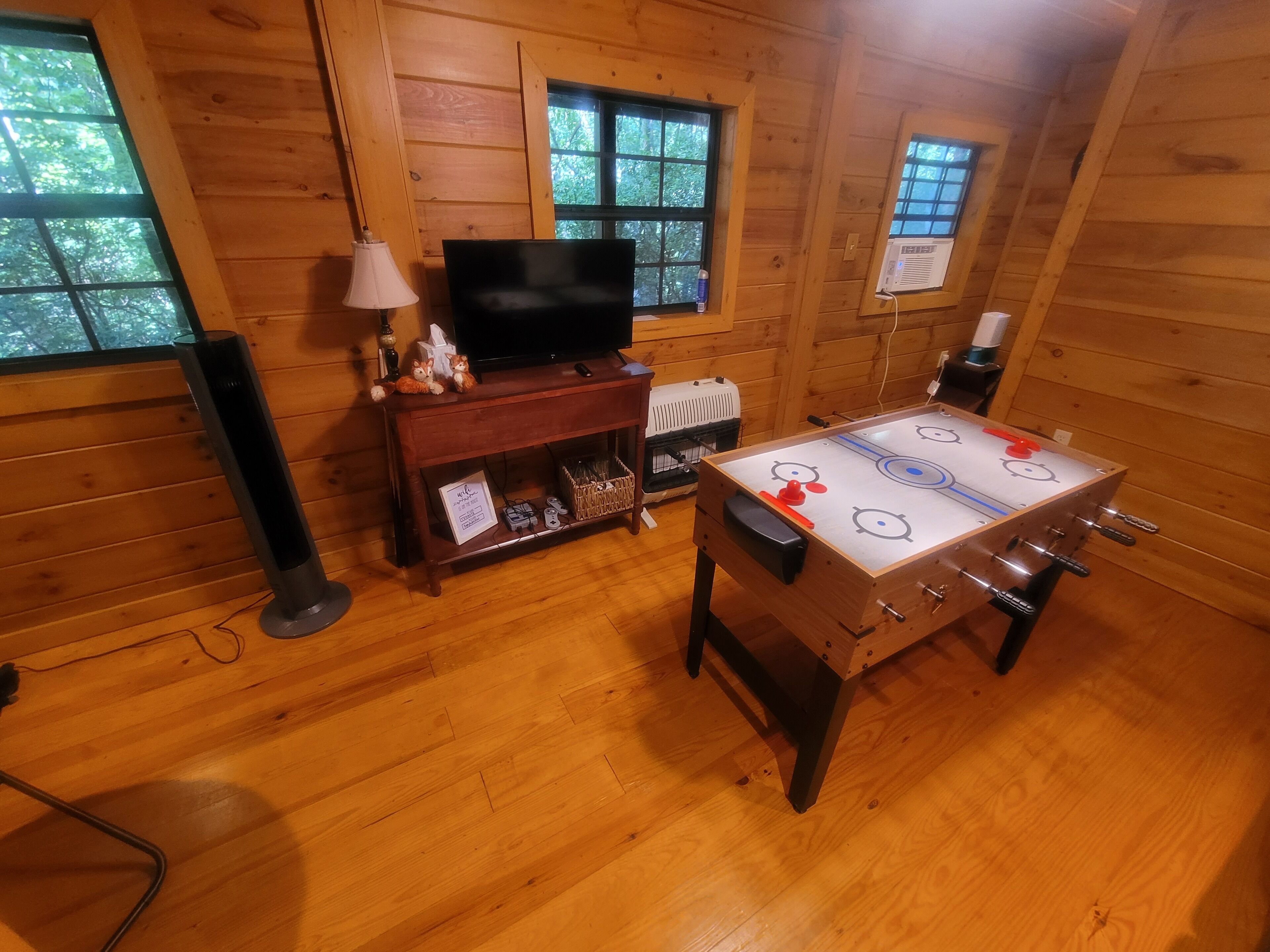 Une pièce chaleureuse aux murs en bois, avec un téléviseur posé sur un meuble en bois, une table de baby-foot et une fenêtre donnant sur les arbres.