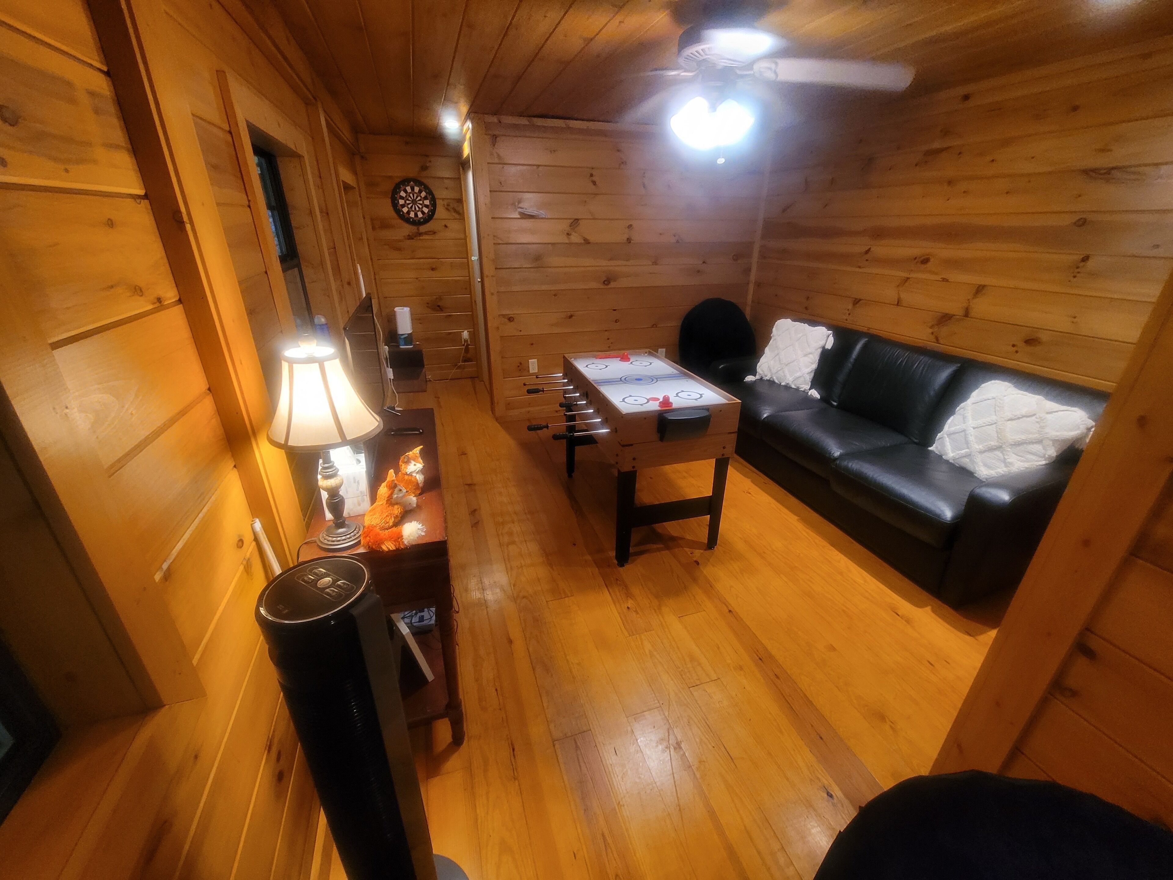 Interior acollidor d'una cabana de fusta, amb sòl de fusta, un sofà de pell negra, una taula petita amb una làmpada i un ventilador de sostre.
