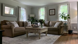 Un salon avec deux canapés beige, une table basse et un tapis. On y trouve des plantes en pot et un tableau encadré accroché au mur.