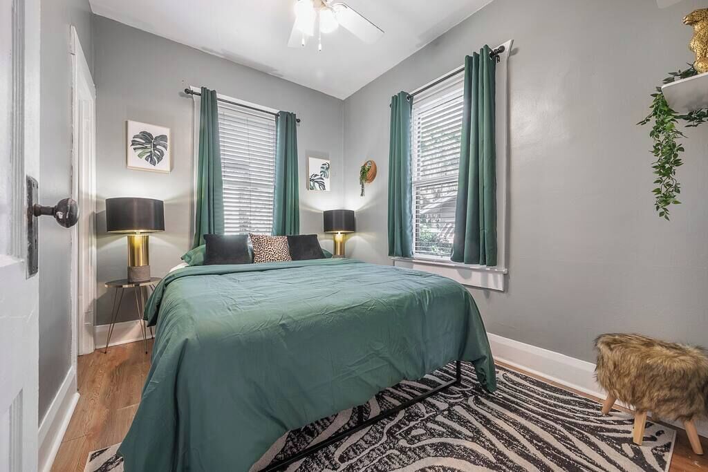 Un dormitorio con una cama, dos lámparas de noche, una ventana con cortinas, una alfombra pequeña y una planta decorativa.