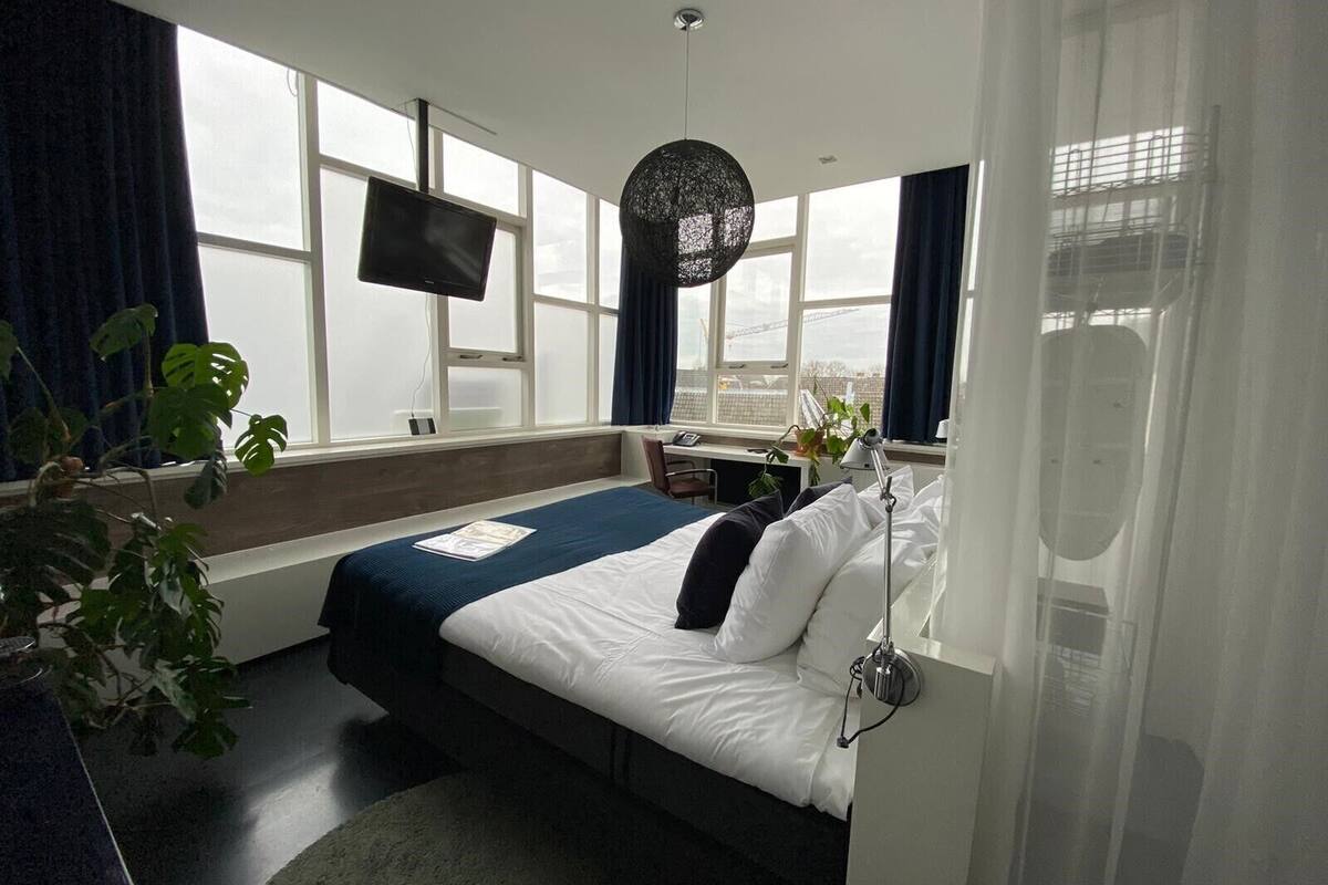 A hotel room with a bed, a desk, a chair, a TV, and large windows.