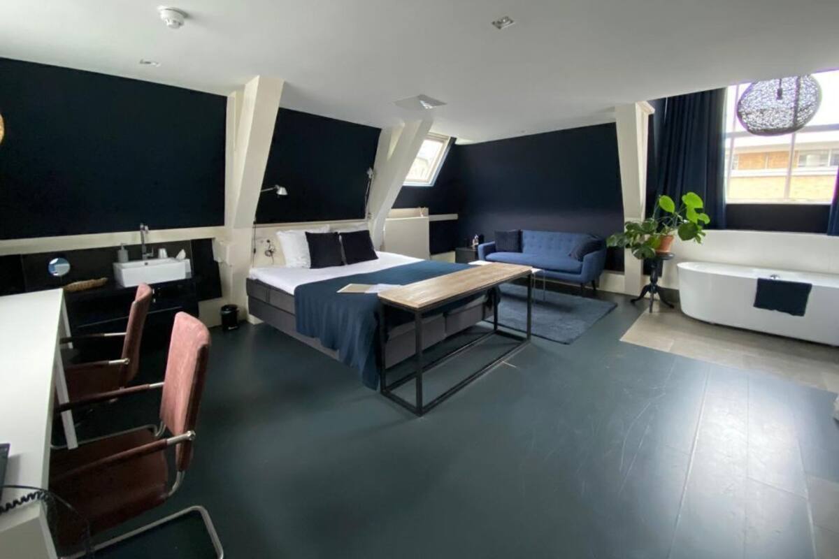 A modern hotel room with a bed, a desk, a chair, a sofa, a bathtub, and a window with curtains.