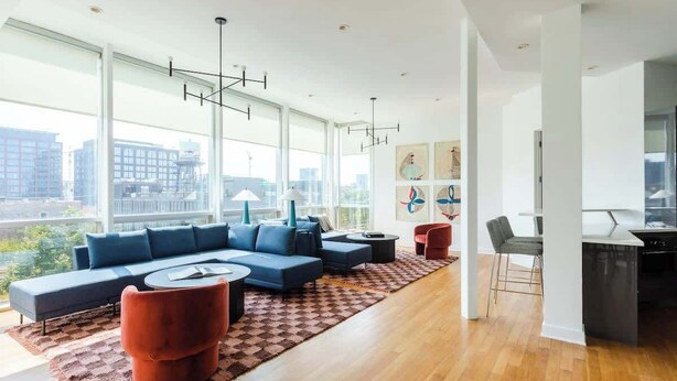 A modern living room with a sectional sofa, a coffee table, and a red chair. There is a kitchen area with a dining table and chairs. The room has large windows with a city view. - Luxury Fulton Market Penthouse Amazing City Views (Chicago)