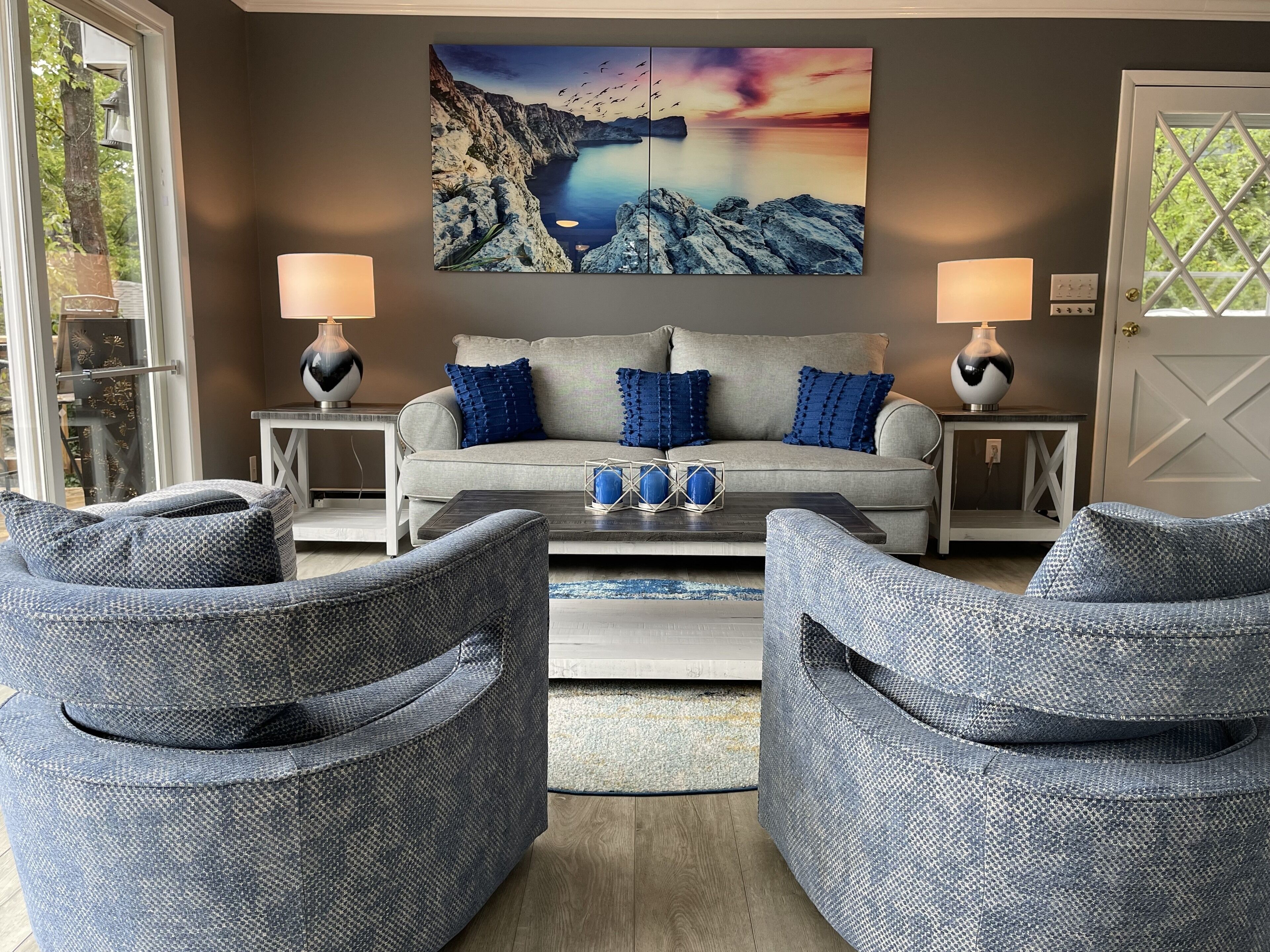 Un salon avec un canapé gris, deux fauteuils assortis, une table basse et un grand tableau représentant un paysage accroché au mur.