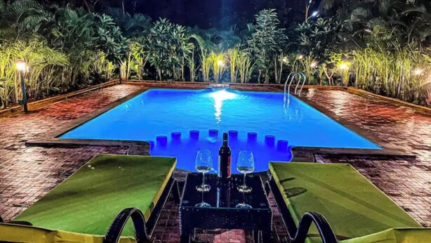 A nighttime pool area with a rectangular pool, two lounge chairs, and a table with a bottle and glasses.