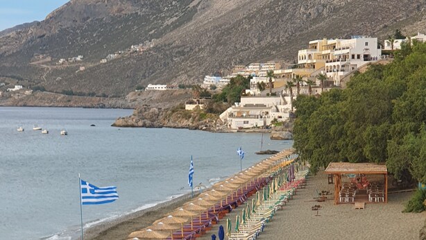A beach with rows of sun loungers and umbrellas, a flag, and a building in the background.
