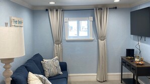 Ein Wohnzimmer mit einem blauen Sofa, einer Lampe, einem Fernseher und einem Fenster mit Vorhängen.