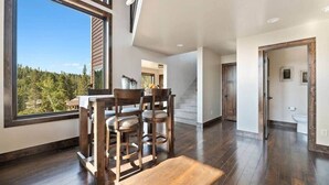 A spacious room with a large window, a dining area with wooden chairs, a staircase, and a door with framed pictures. - Altitude Adjustment (Lead)