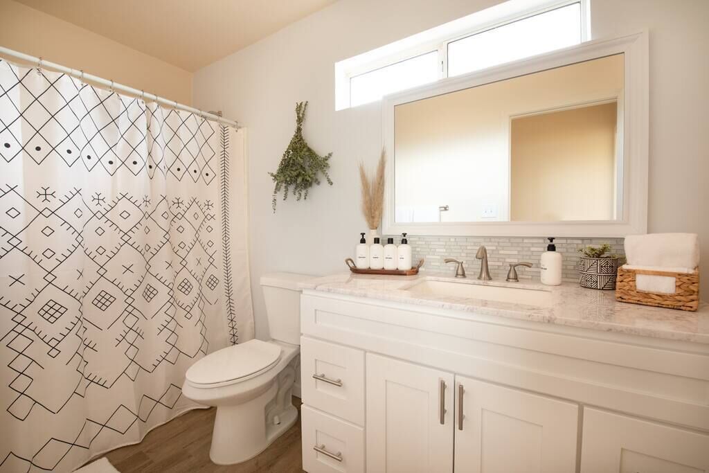Un baño con cortina de ducha estampada, un inodoro, un lavabo con espejo y un mueble con artículos de aseo.