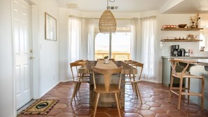 Comedor con mesa y sillas de madera, lámpara colgante y vista a través de una ventana con cortinas.