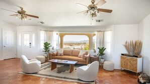 Un salón amplio con un ventanal grande, un sofá color beige, dos sillones blancos, una mesa de centro y un ventilador de techo.