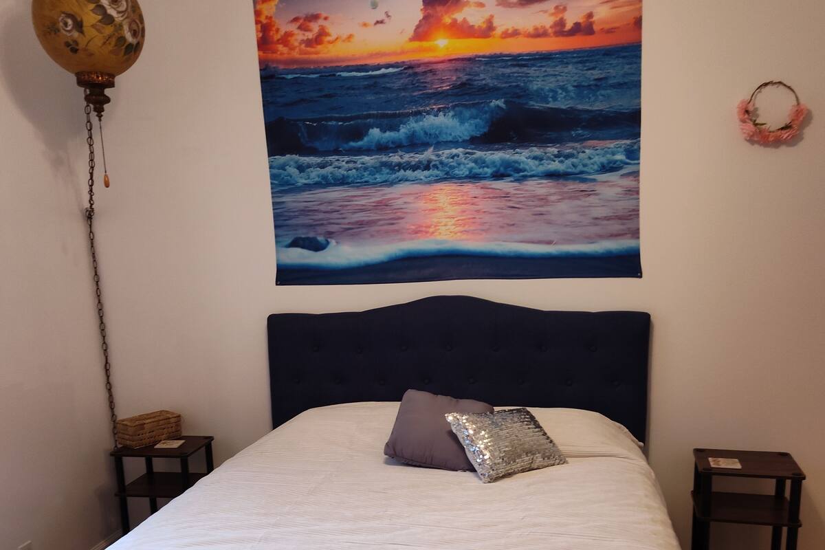 A bedroom with a bed, a wall-mounted tapestry of a sunset over the ocean, a hanging lamp, and a small nightstand.