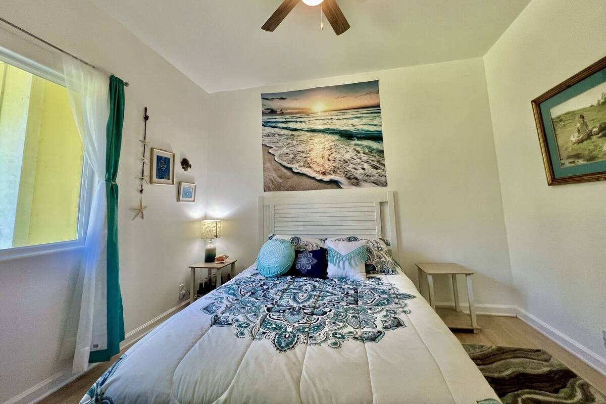 A bedroom with a bed, a ceiling fan, a wall-mounted painting of a beach scene, and a window with curtains.